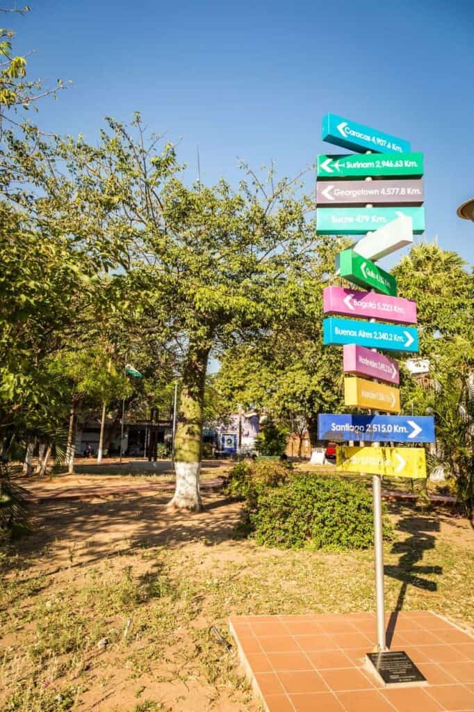 A multi-colored signpost in Santa Cruz, Bolivia points toward major cities around the world, including Caracas, Buenos Aires, Bogotá, and Santiago, with distances in kilometers. The sign stands on a tiled base in a sunny park surrounded by trees and greenery.