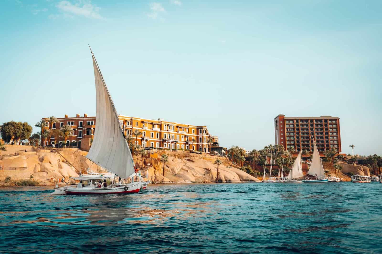A white felucca sailboat glides along the Nile River in Aswan, Egypt, with the iconic Old Cataract Hotel and modern hotel buildings rising behind rocky shores.