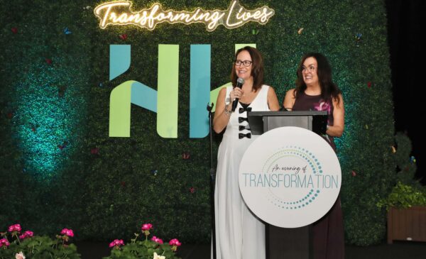 Two women stand on a stage at a podium labeled “An Evening of Transformation.” One speaks into a microphone. Behind them is a green backdrop with "Transforming Lives" in neon and geometric shapes. Pink flowers are in the foreground.