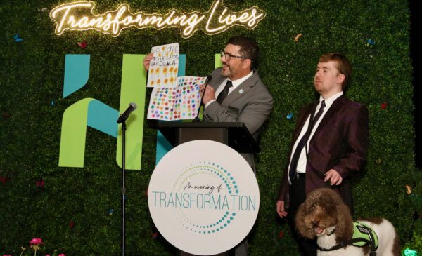 Two men stand on stage at a podium with a “Transforming Lives” neon sign. One man holds a colorful poster, while the other stands beside him with a service dog. The backdrop is green foliage with event branding.