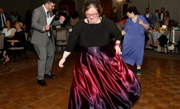 A woman in a shiny purple skirt dances on a wooden floor at a formal event, with other people in suits and dresses dancing and sitting at tables in the background.