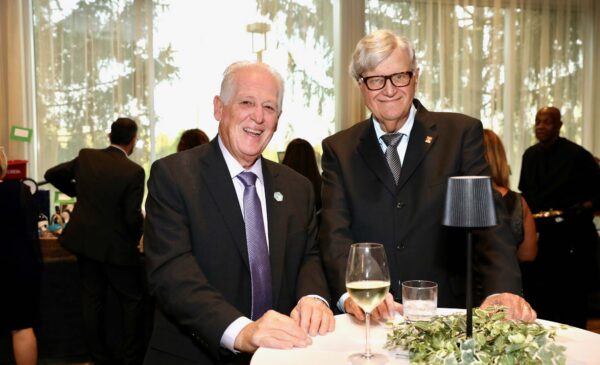 Two older men in suits smile while standing at a round table with wine glasses and a small lamp. People and large windows with curtains are in the background, suggesting a formal event or gathering.