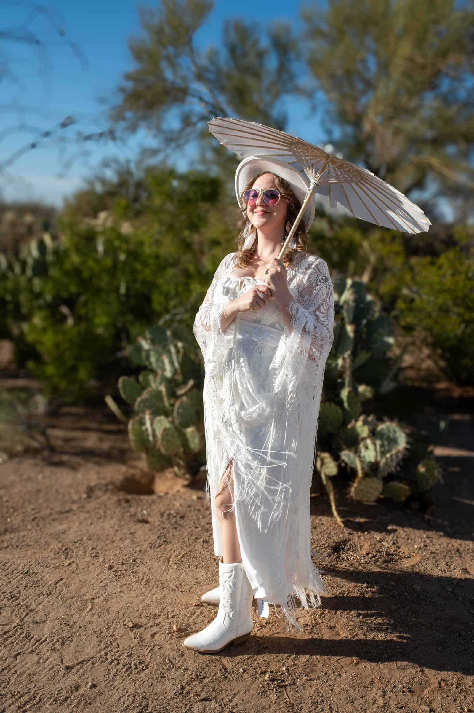 A woman dressed in a flowing white gown with sunglasses, holding a parasol, standing in a desert landscape with cacti and trees.