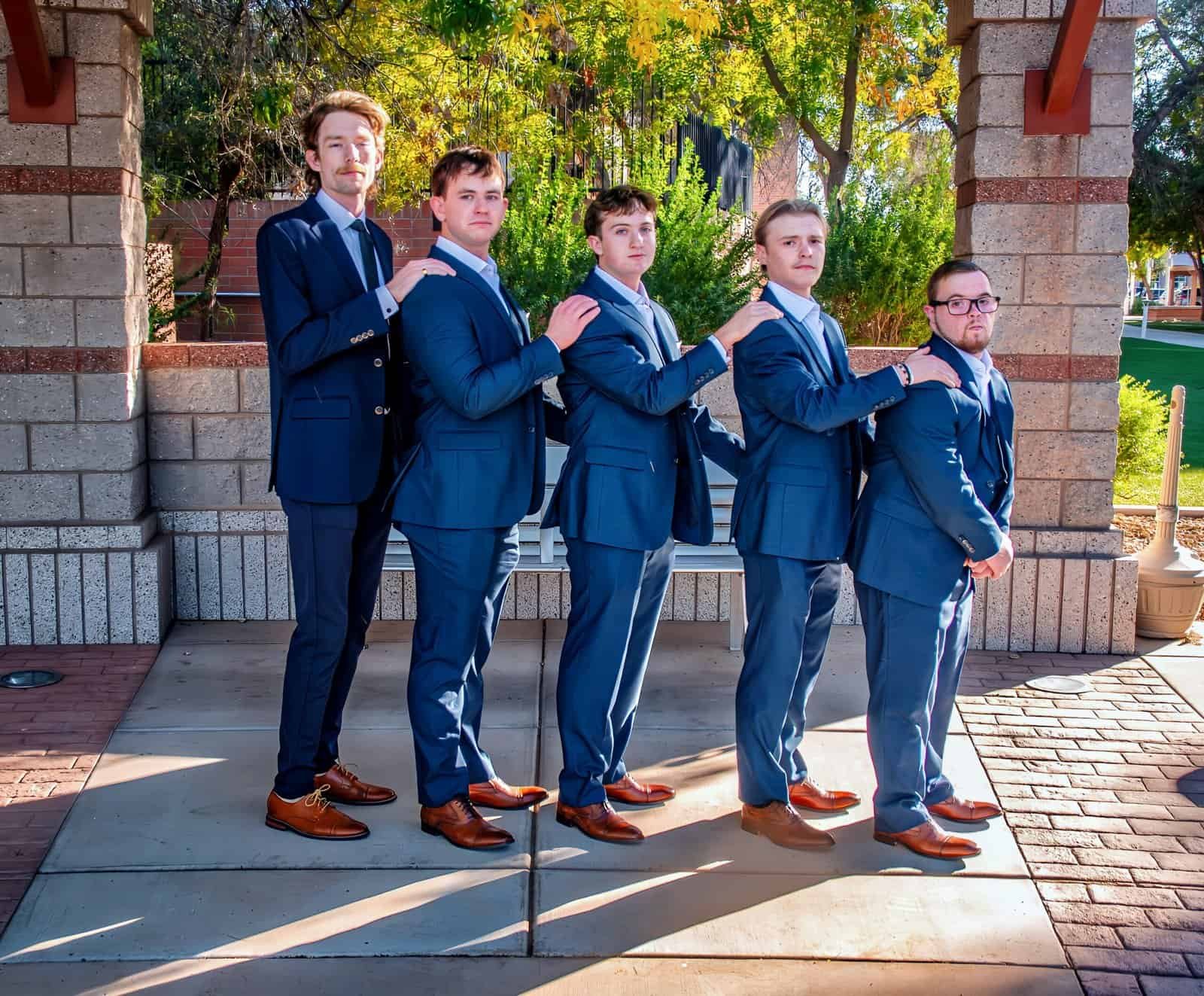 A group of five groomsmen dressed in matching suits, standing in a line outdoors during a wedding celebration.