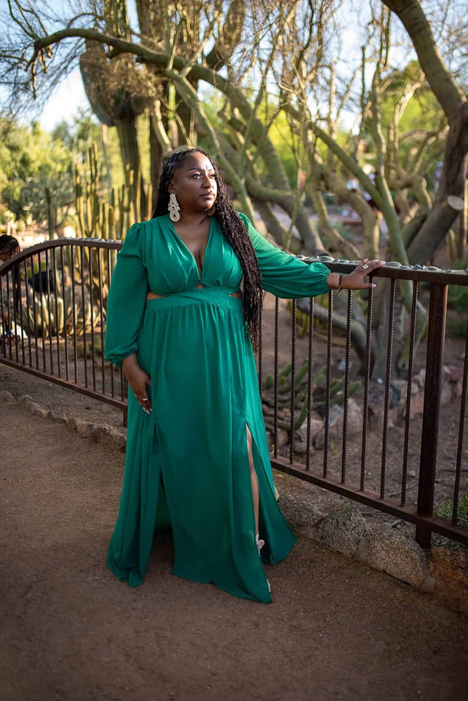 A woman in a vibrant green dress stands gracefully by a park railing, surrounded by trees and nature, capturing a serene moment.