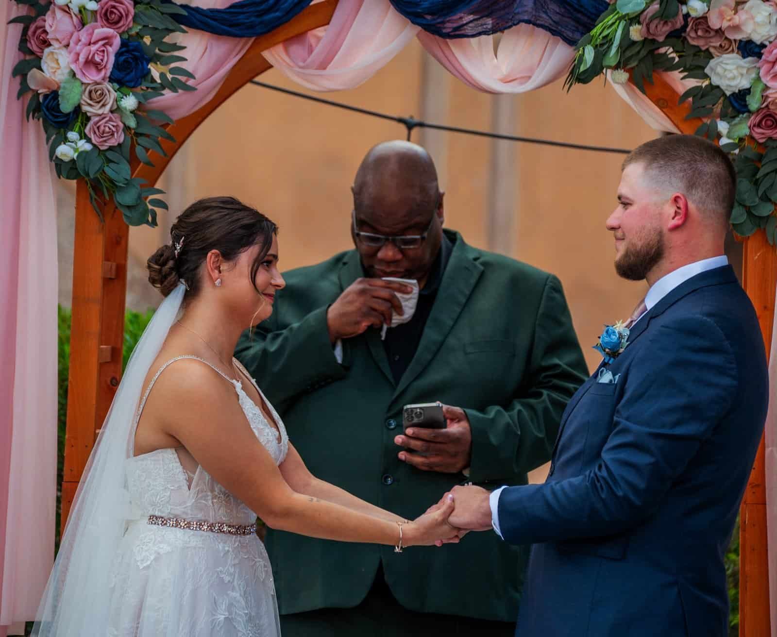 Beautiful wedding moment with bride and groom holding hands under a decorated floral arch during their vows.