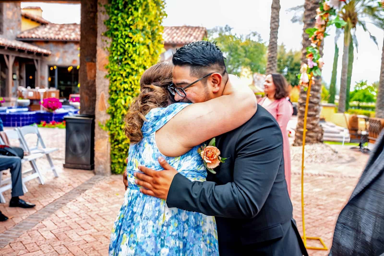 A joyful couple shares a heartfelt hug at their wedding celebration in a scenic outdoor setting with guests and decorative elements.