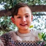 Child smiling outdoors in a natural setting with trees and leaves.