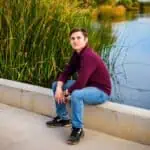 Young man sitting by a peaceful lake with tall grass in the background.