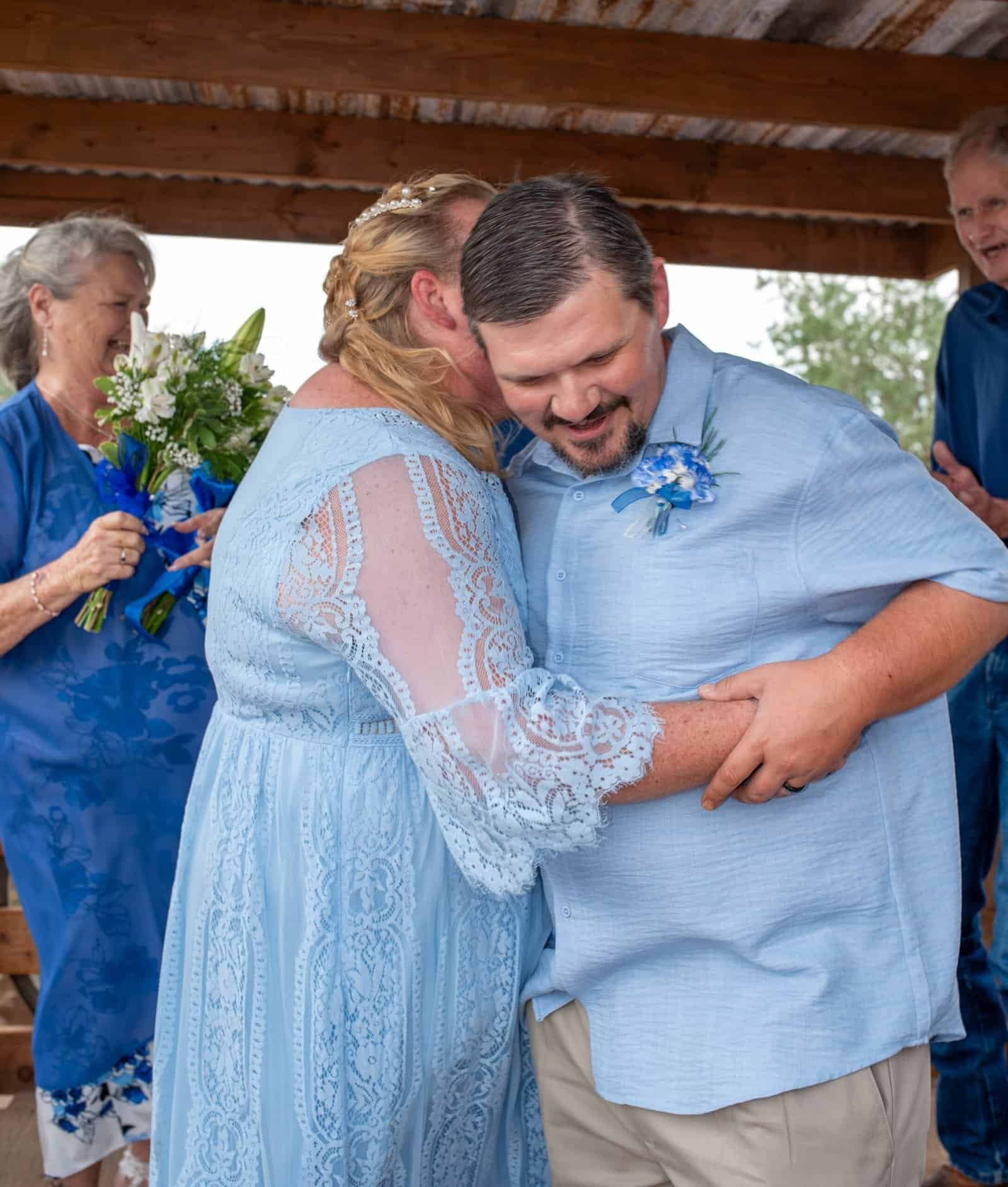 A bride and groom share a dance during their wedding celebration, surrounded by family and friends in a rustic outdoor setting.