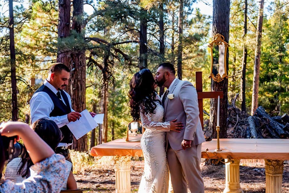 A couple shares a kiss during their outdoor wedding ceremony in a scenic forest surrounded by tall trees.