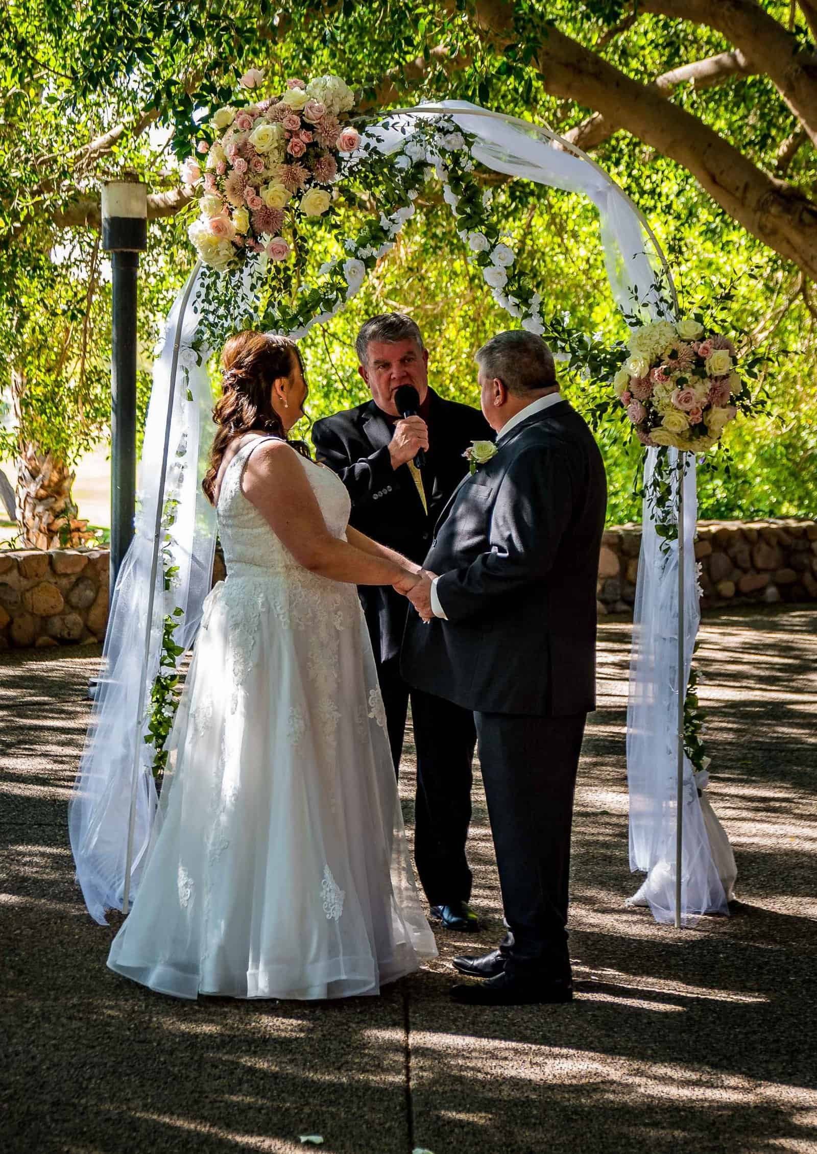 Beautiful outdoor wedding with floral arch, couple exchanging vows, and officiant present.