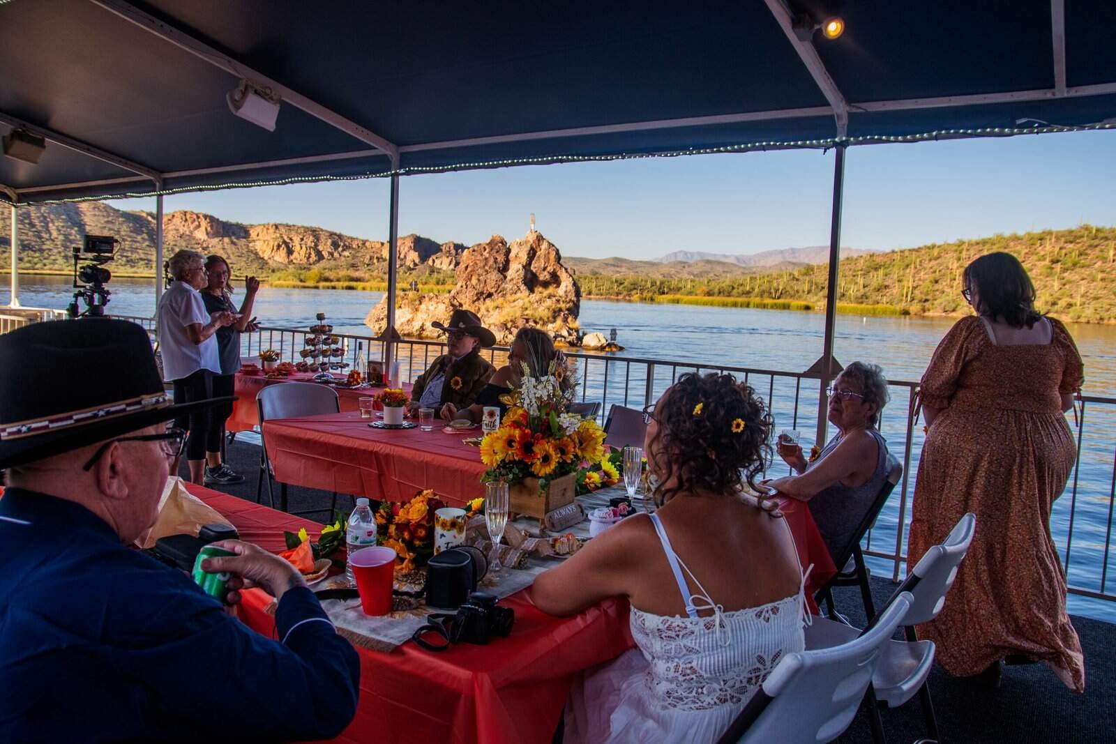 A group of people celebrating a wedding on a boat with a beautiful lake and mountain scenery, featuring a decorated table and joyful atmosphere.