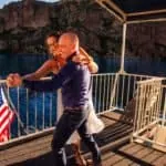 Happy couple dancing on boat deck with scenic water and cliffs in background.