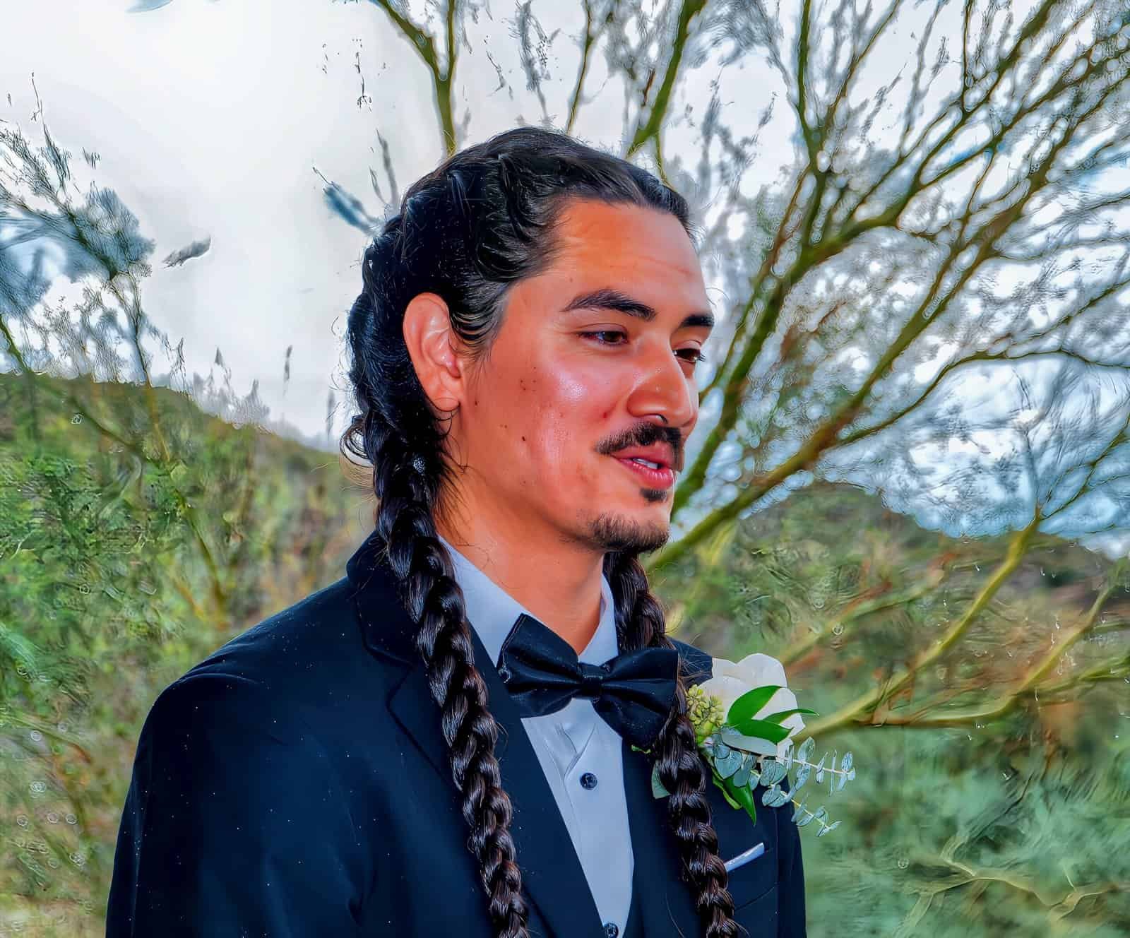 A groom dressed in a suit with a bow tie, standing outdoors with trees and nature in the background.