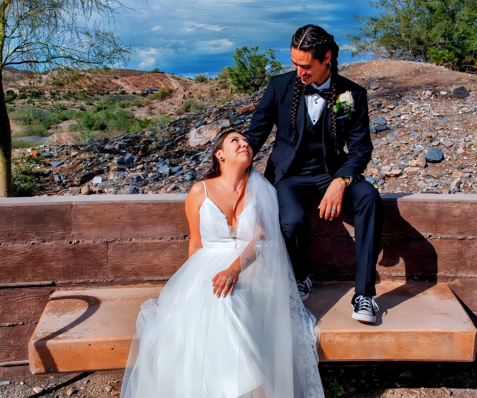 A happy bride and groom sitting on a wooden bench outdoors, surrounded by desert scenery and blue sky.