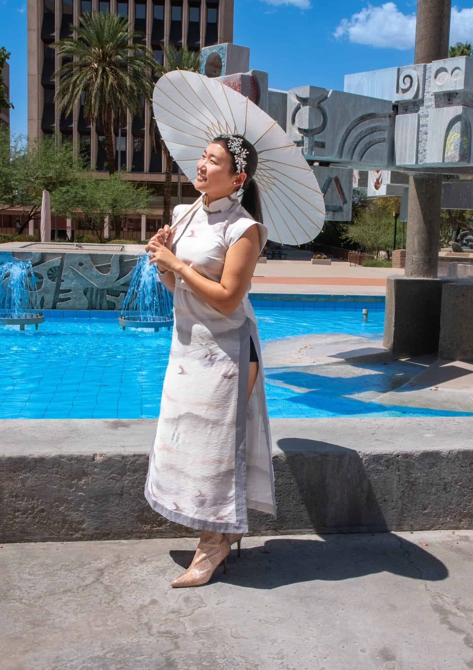 A woman dressed in traditional attire holding a parasol near a fountain in an urban park setting.