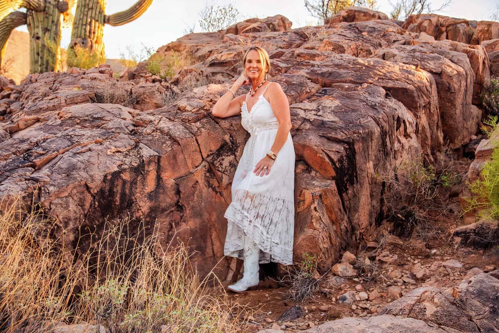 A woman in a white dress poses on a rocky desert landscape with cacti and natural rock formations, capturing a joyful moment outdoors.