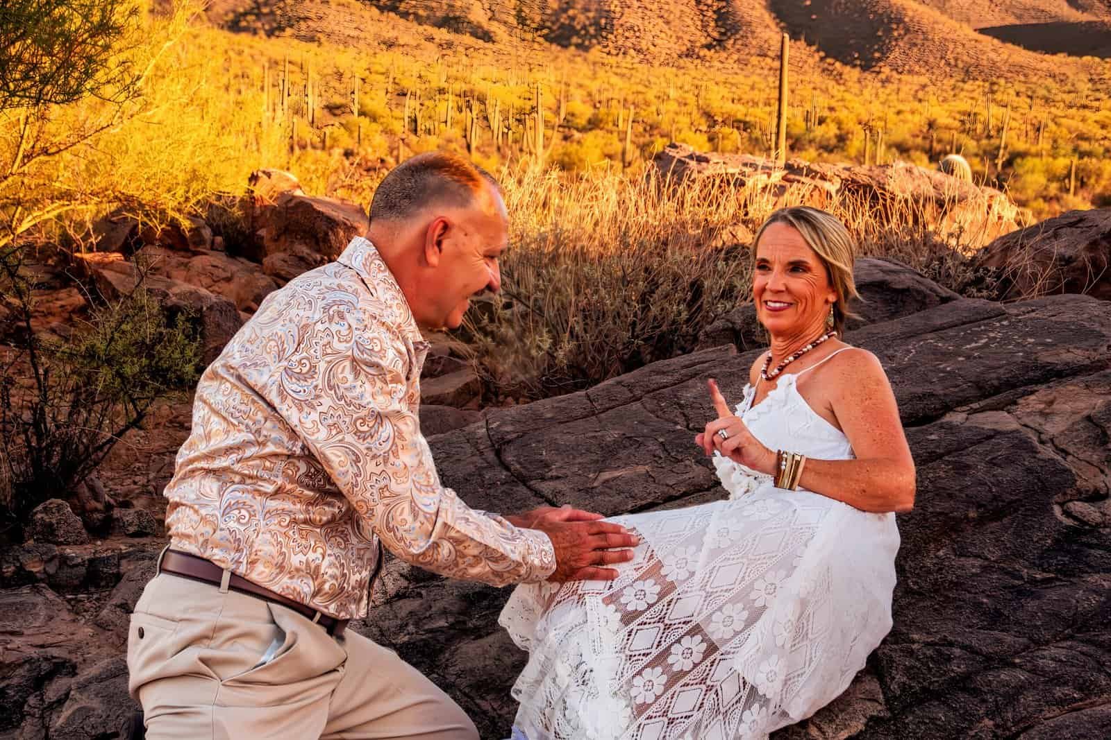 A romantic wedding proposal scene outdoors during sunset, featuring a man proposing to a woman on rocky terrain.