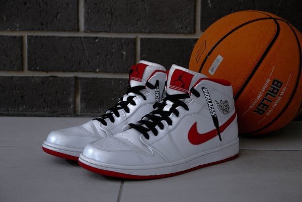 1. Pair of white and red Nike high-top sneakers with black laces next to a basketball on a grey floor against a brick wall.