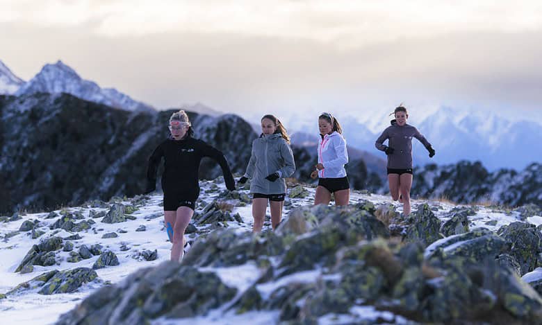 Mountain trail runners in snowy terrain with rugged rocks and snow-capped peaks in the background, showcasing outdoor running and adventure sports.