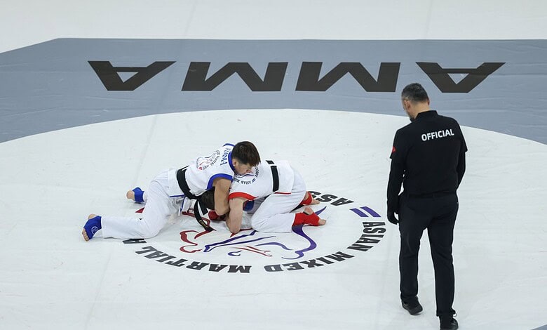 An official overseeing a judo match on a professional mat, with two athletes engaged in a grappling technique, highlighting competitive martial arts and sports officiating.
