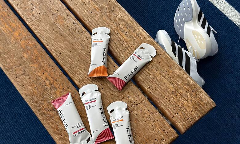 Assorted sports nutrition gels and energy chews placed on a wooden bench next to running shoes on a blue track surface.