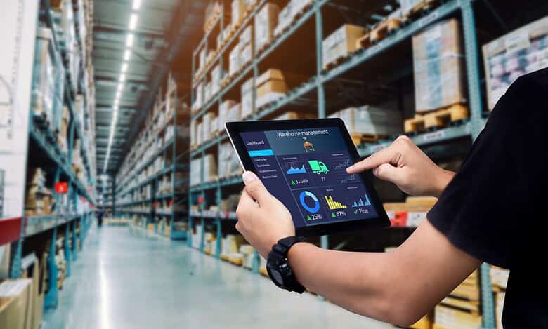 Warehouse worker using a tablet for inventory management among shelves filled with boxes and supplies. Modern logistics and stock control in a large storage facility.