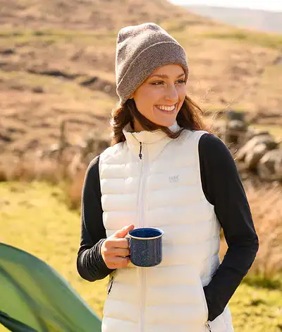 Outdoor woman smiling and holding a blue mug in a scenic landscape with hills and grass.