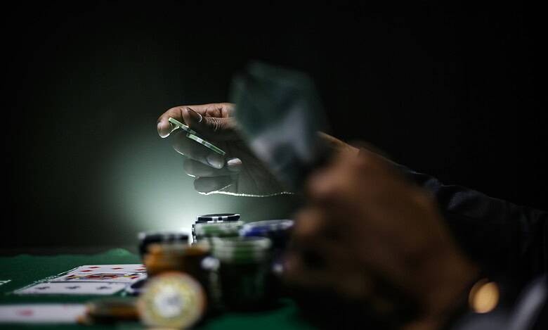 Close-up of a person using a smartphone for online sports betting, with poker chips and playing cards on a green table, highlighting sports gambling and betting activities.