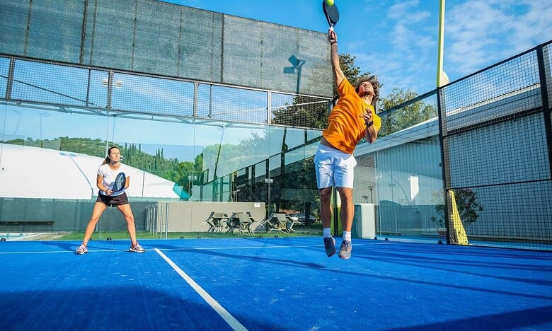 Two players competing in a padel tennis match on an outdoor court with a blue surface and glass walls under a clear sky.