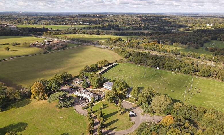Aerial view of sports fields, lush greenery, and surrounding countryside under a partly cloudy sky.