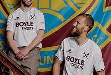 Two West Ham United players in the new 2026/27 away kit, standing on a staircase with a club banner in the background, smiling and engaging.