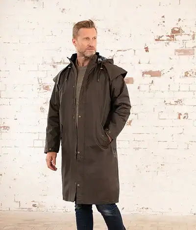 Man wearing a full-length waterproof rain coat in dark brown, standing outdoors against a white brick wall, showcasing the coat's durability and style.