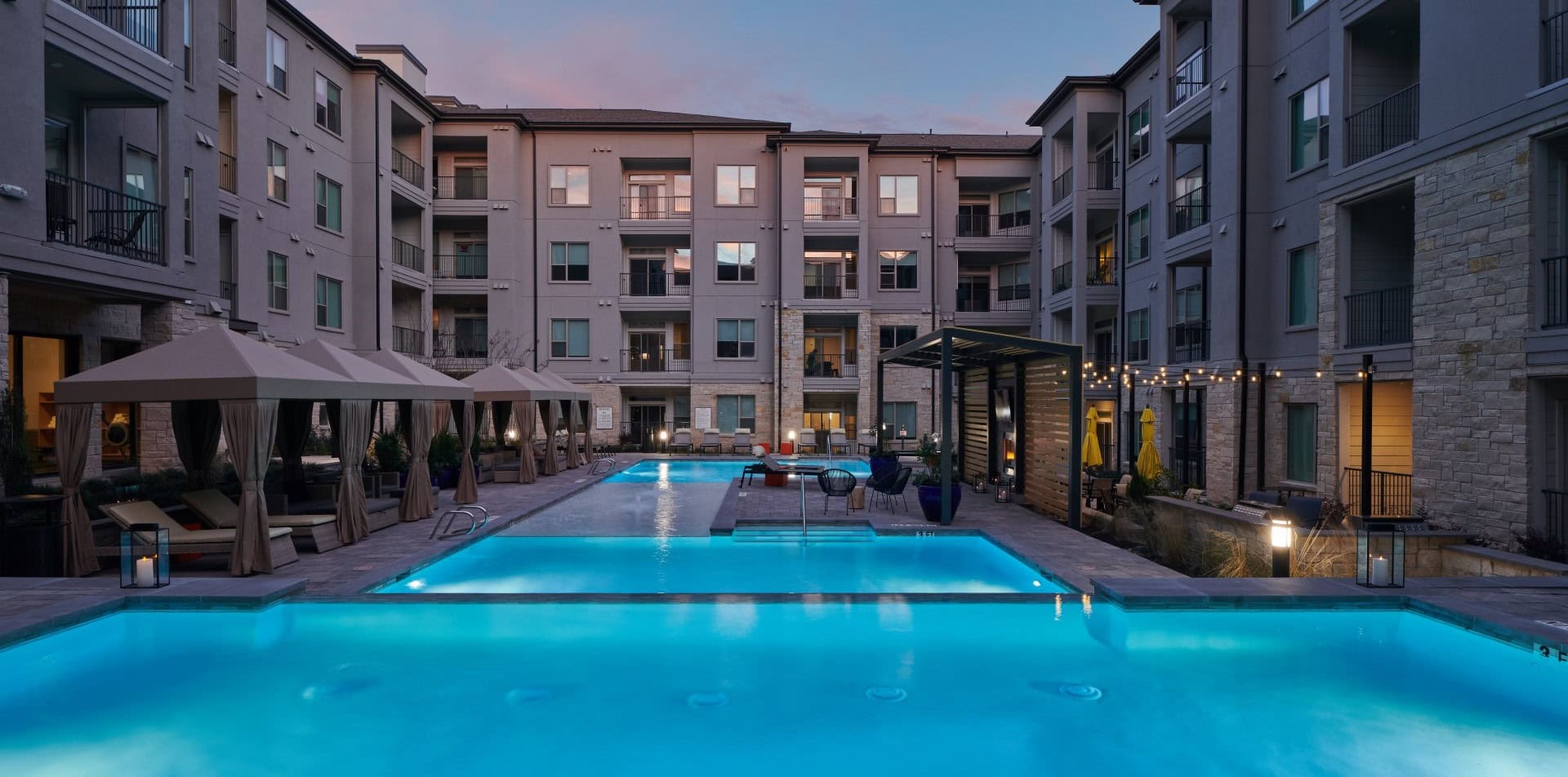 Windsor Burnet is a modern apartment complex at dusk with illuminated swimming pools, cabanas, string lights, and balconies overlooking the courtyard, creating a relaxing and inviting atmosphere.