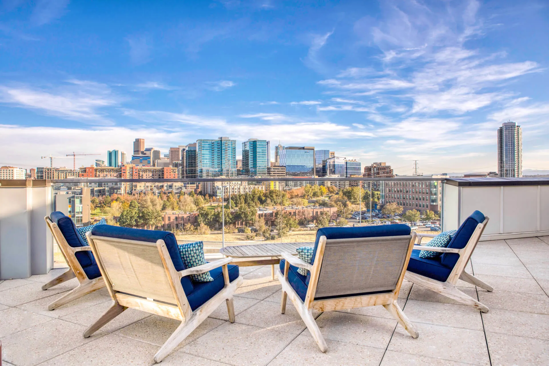 Four cushioned patio chairs surround a small table on a rooftop terrace at Centric LoHi, featuring a glass railing and overlooking a city skyline with modern buildings under a blue sky dotted with wispy clouds.