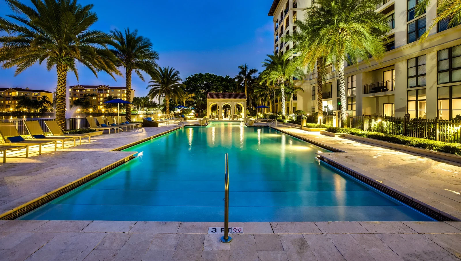 A luxurious outdoor swimming pool at Windsor at Doral is surrounded by palm trees, lounge chairs, and elegant buildings, illuminated at dusk with shimmering reflections. A handrail and 