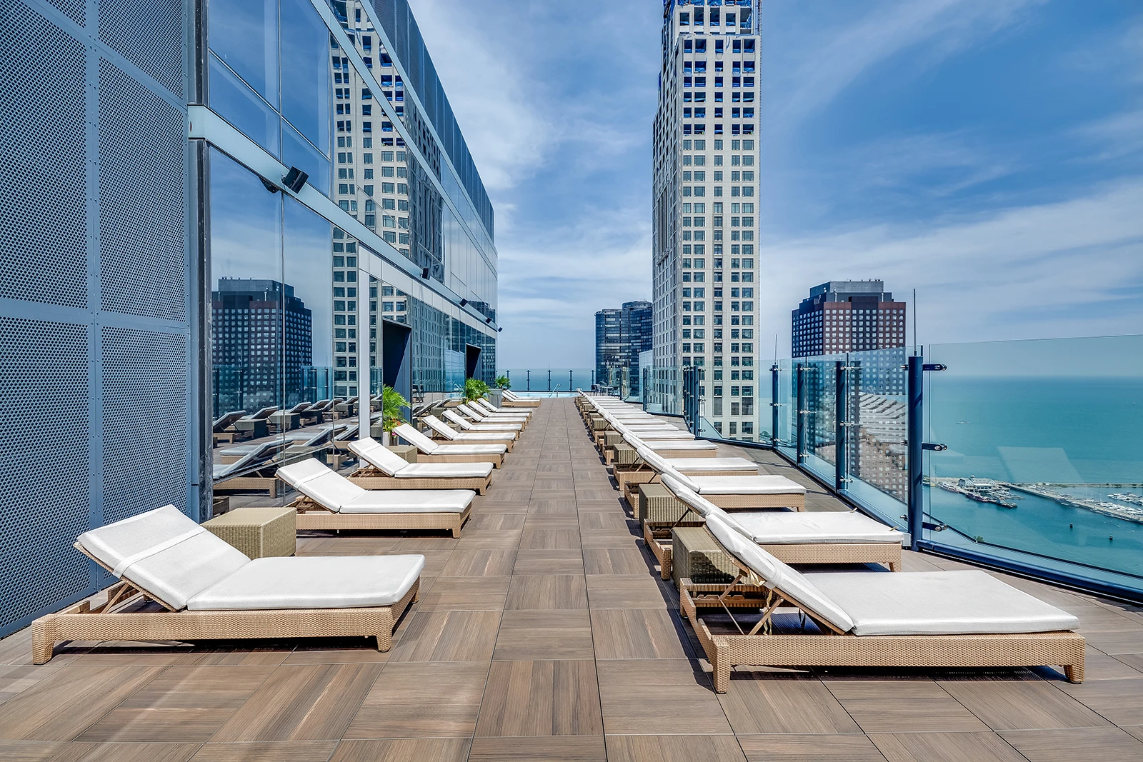 A rooftop deck with rows of white cushioned lounge chairs faces glass railings, inviting you to savor each moment while overlooking tall city buildings and a vast body of water beneath a blue sky.