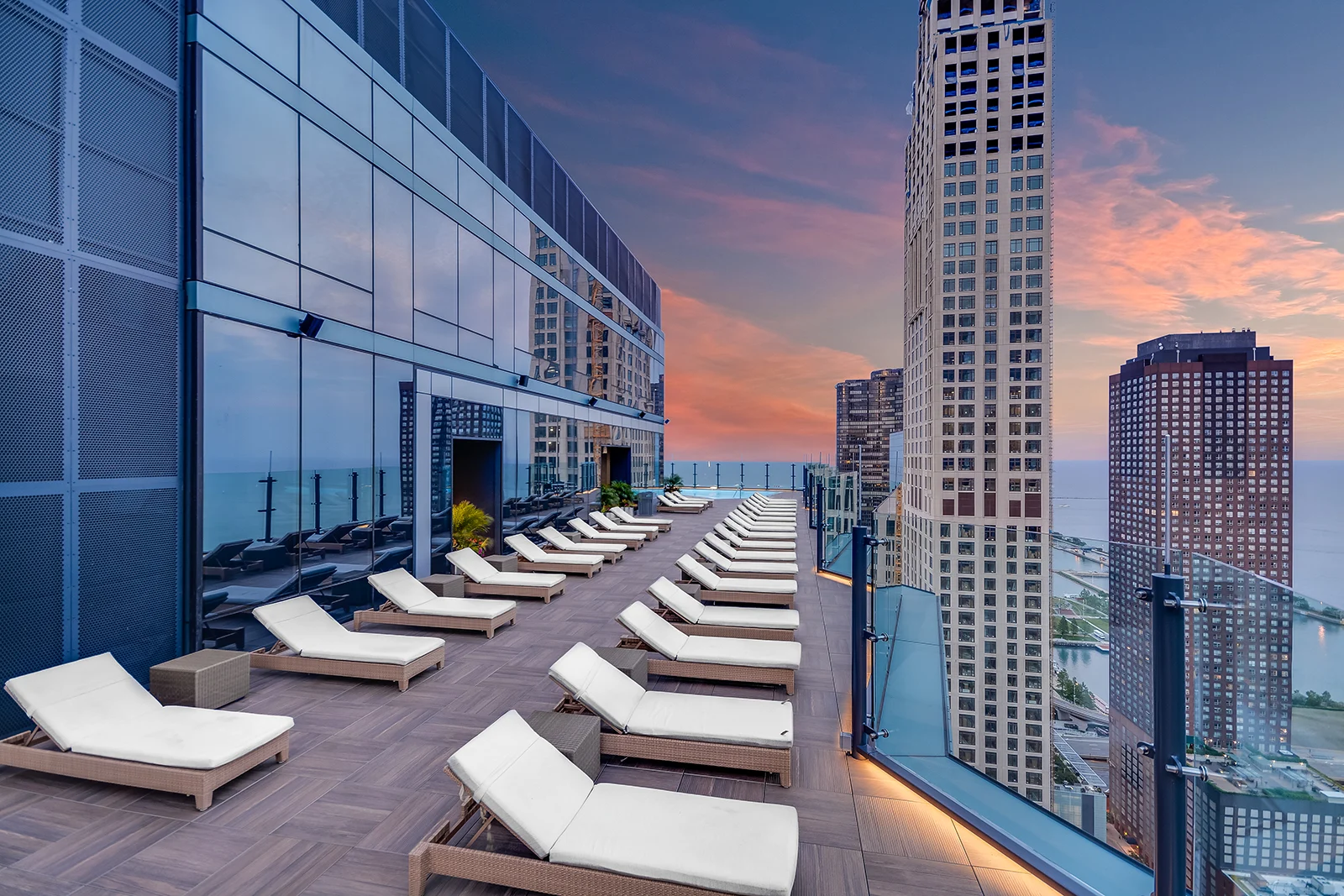 A modern rooftop terrace captures the moment with rows of white lounge chairs overlooking tall city buildings and a lake at sunset, as glass railings and reflective windows enhance the urban view.
