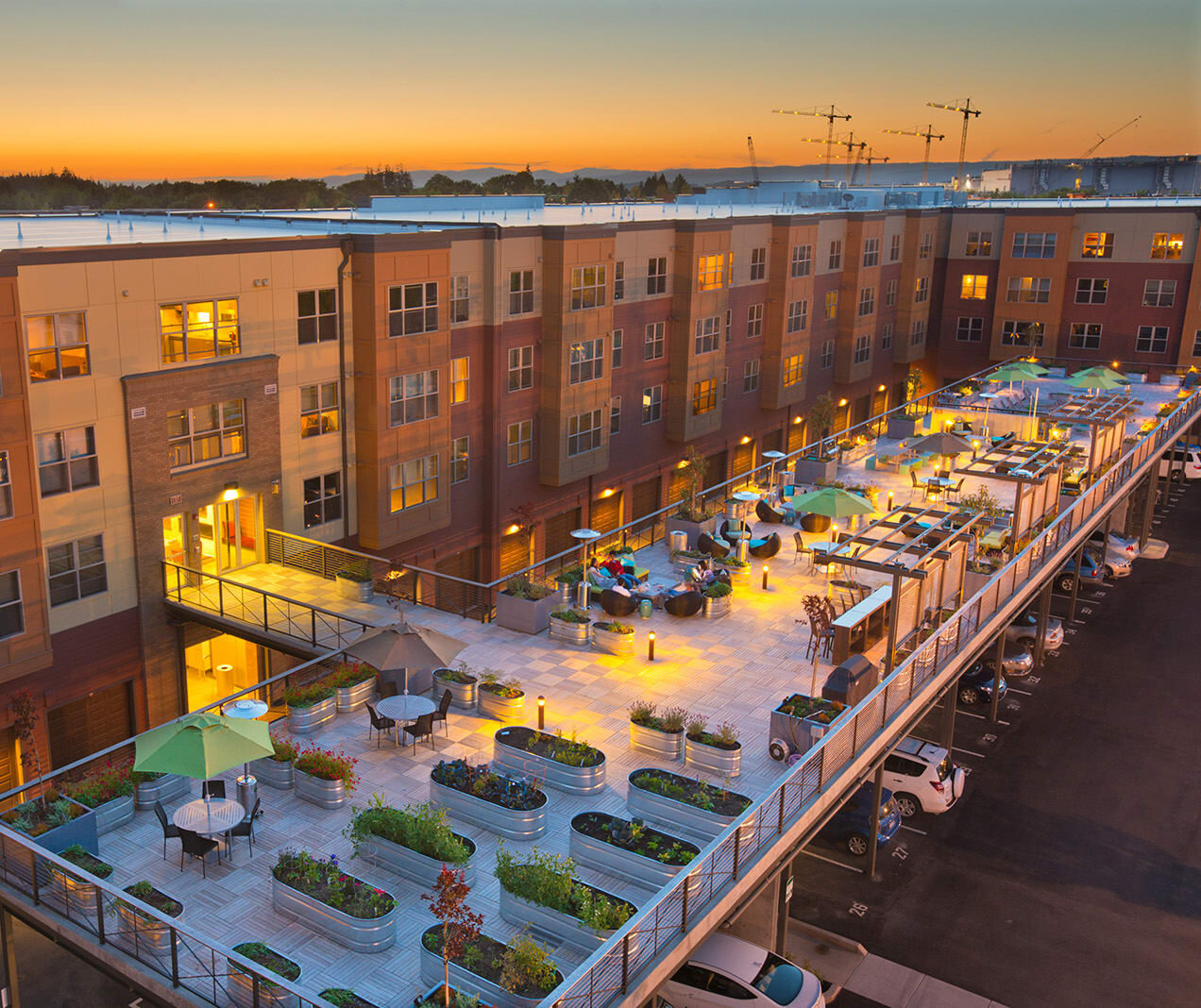 A rooftop terrace at Platform 14 features seating areas, plants, and lights atop a modern apartment building at sunset. Cars are parked below, with construction cranes visible in the background.