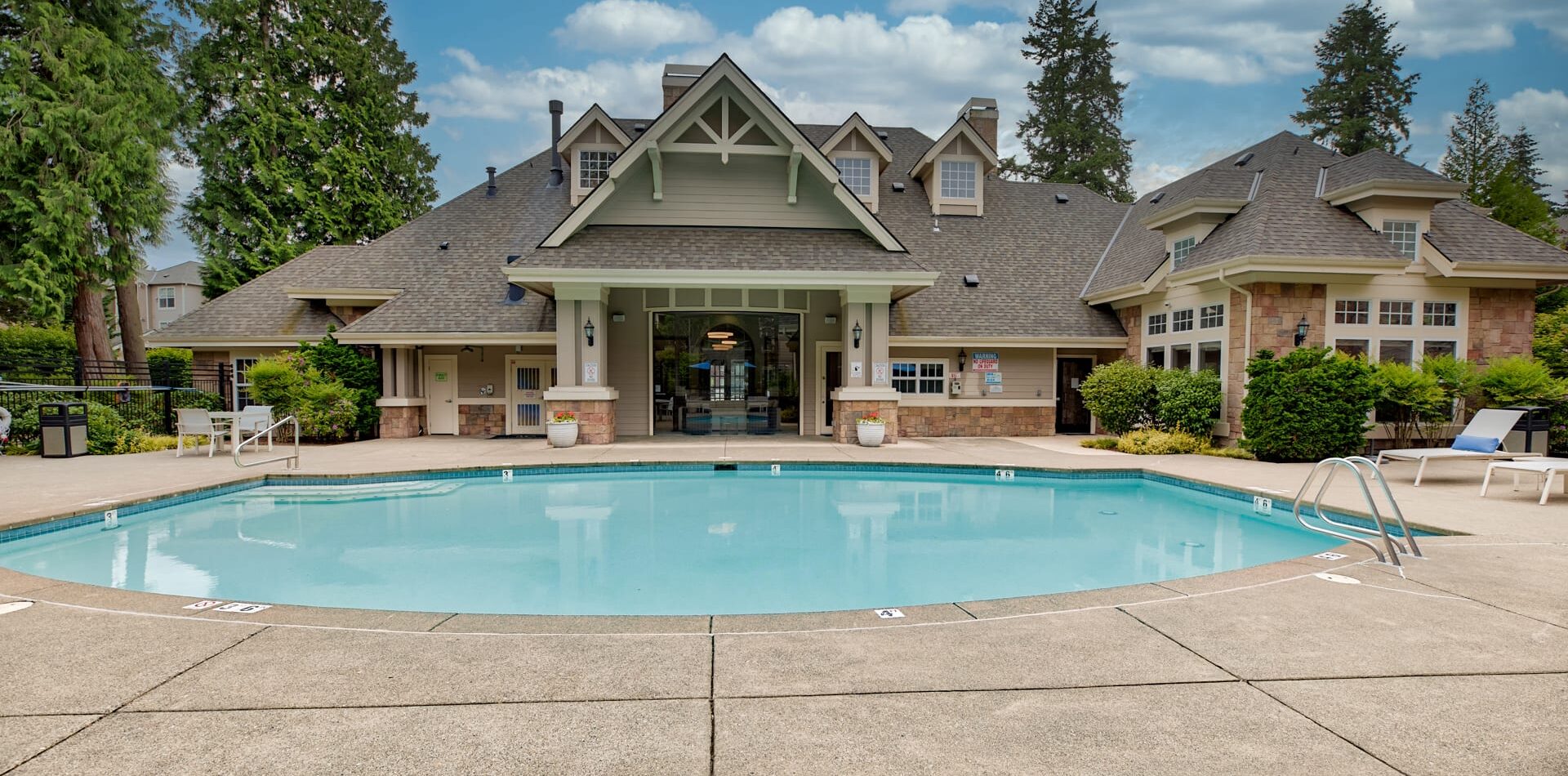 A clear outdoor swimming pool with a concrete deck sits before the lodge-style Reflections by Windsor building, surrounded by trees under a partly cloudy sky. Lounge chairs and greenery line the peaceful pool area.