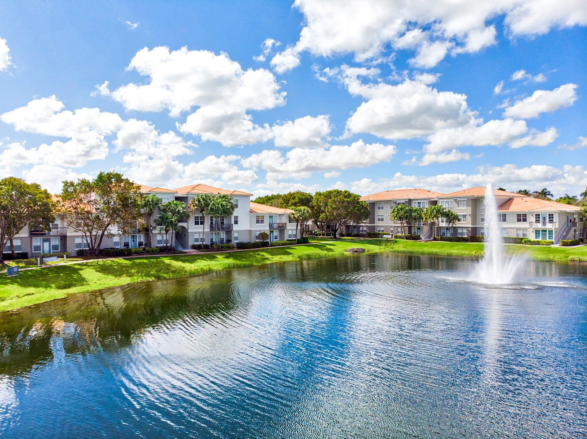 A scenic pond with a fountain is surrounded by green grass, palm trees, and the elegant white apartment buildings of Windsor at Miramar under a blue sky with scattered clouds.