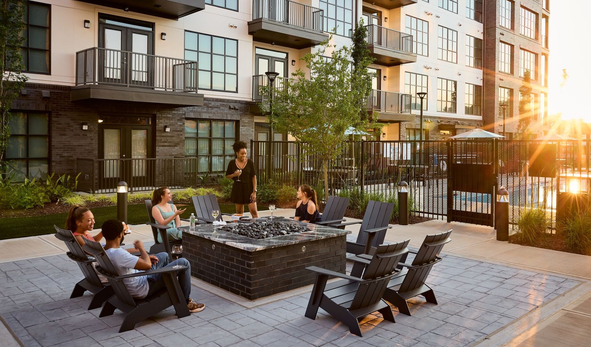 Five people sit and chat around an outdoor fire pit at 565 Hank's modern apartment courtyard, with the sun setting in the background and a pool visible behind a fence.