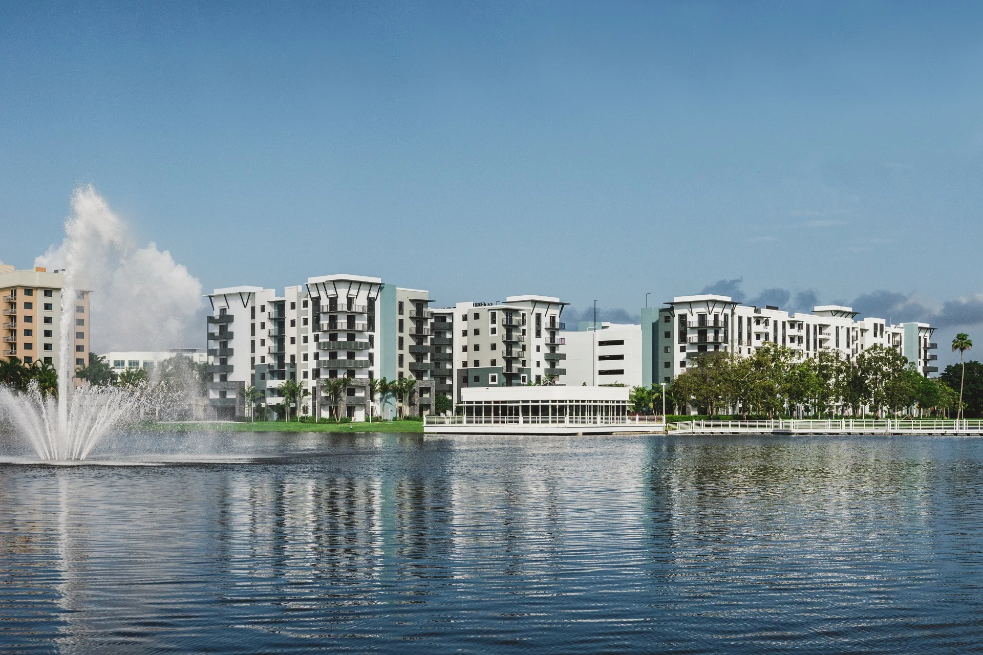 Windsor Cornerstone is a modern apartment complex with several mid-rise buildings standing behind a lake; a fountain sprays water on the left, and palm trees line the shore under a clear blue sky.