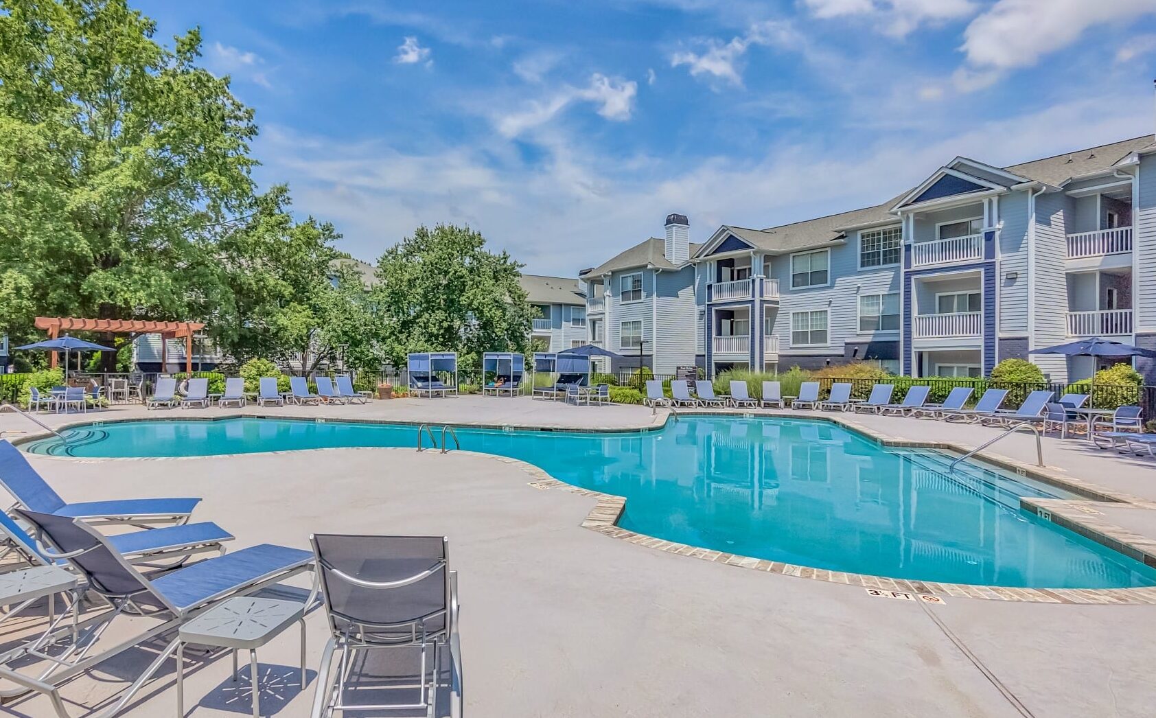 Bright outdoor swimming pool at Windsor Addison Park, surrounded by lounge chairs and tables, with light blue apartment buildings and balconies in the background under a partly cloudy sky.