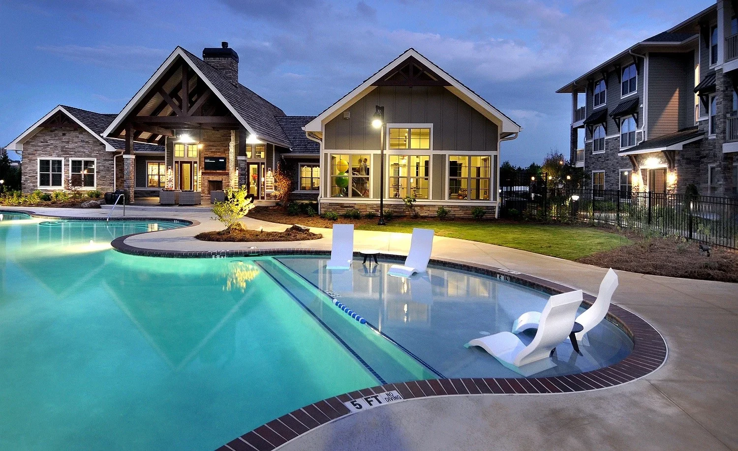 Modern pool area at dusk with submerged lounge chairs in shallow water, a large clubhouse with tall windows and peaked roof, and an apartment building in the background. The lights create a welcoming, relaxing atmosphere.