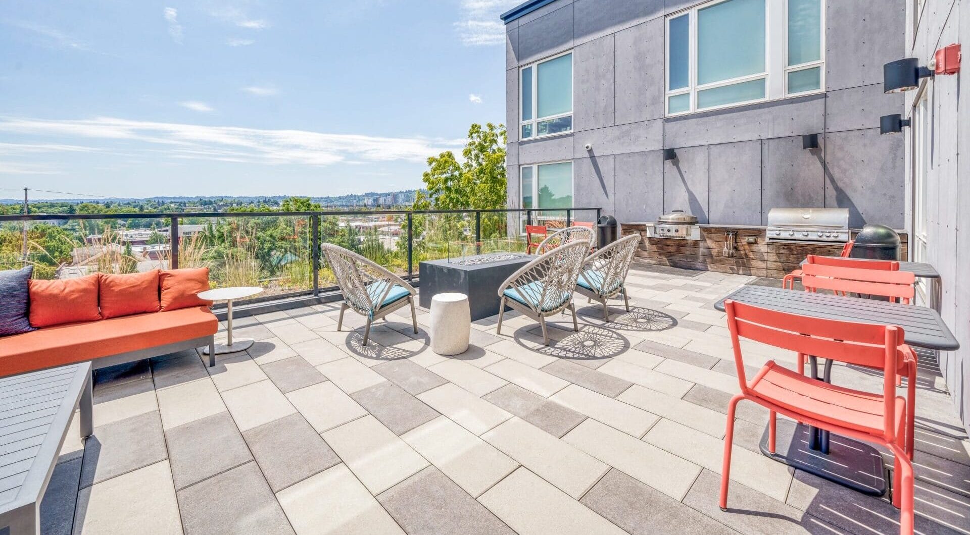 A modern rooftop patio at Windsor Buckman features lounge seating, wicker chairs, red dining chairs, tables, a grill area, and a scenic city view under a partly cloudy sky.