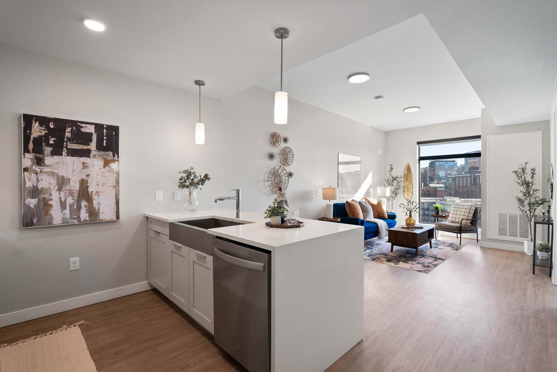 Modern open-concept apartment at Windsor 3000 Huron with a white kitchen island and sink, stainless steel appliances, contemporary decor, and a bright living area featuring large windows overlooking a stunning cityscape.