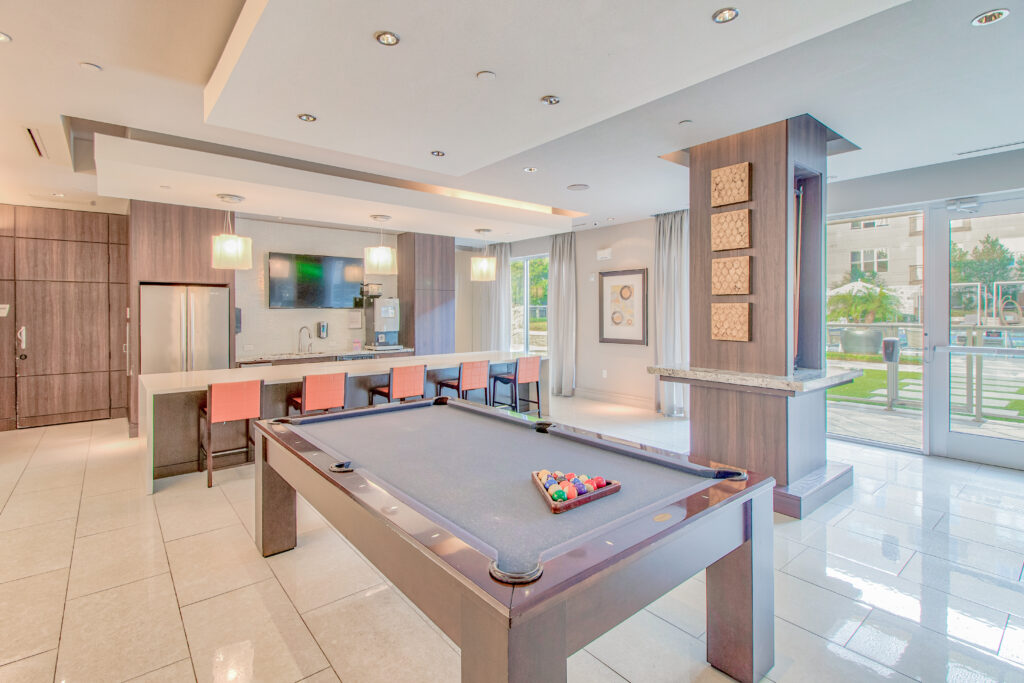 A modern lounge with a pool table in the center, a bar area with four orange chairs, a wall-mounted TV, and large windows showing an outdoor pool and greenery. The room features sleek, contemporary decor.