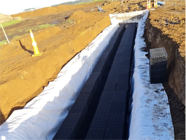 Stormwater drainage channel construction with geotextile fabric and black plastic drainage pipes, civil engineering site in progress.